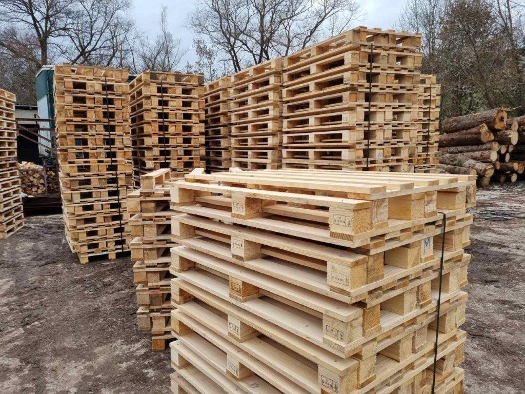 Piramida od drvenih paleta na površini prekrivenoj šljunkom sa drvećem i prenosivim toaletom u pozadini.